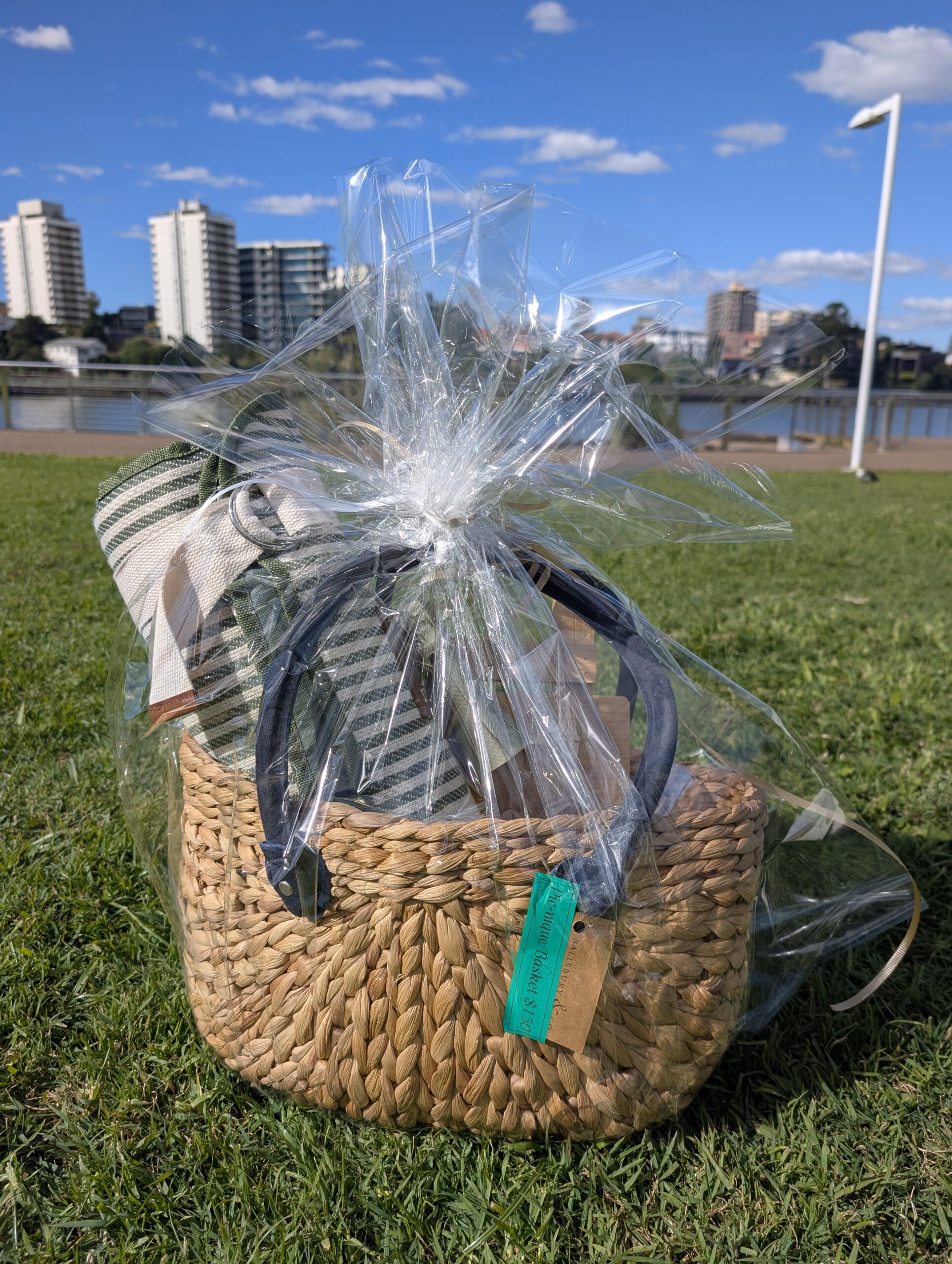 A beautiful Pic-nique by Medley basket wrapped in cellophane, sitting on green grass with a city skyline in the background.